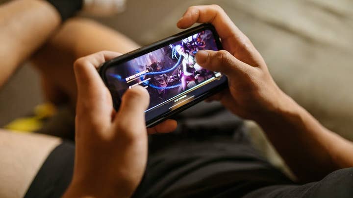 Two hands hold a phone sideways and play a game on it with the camera viewing it from an angle near the person's shoulder. Their legs and body are visible behind the phone, somewhat blurred and out of focus